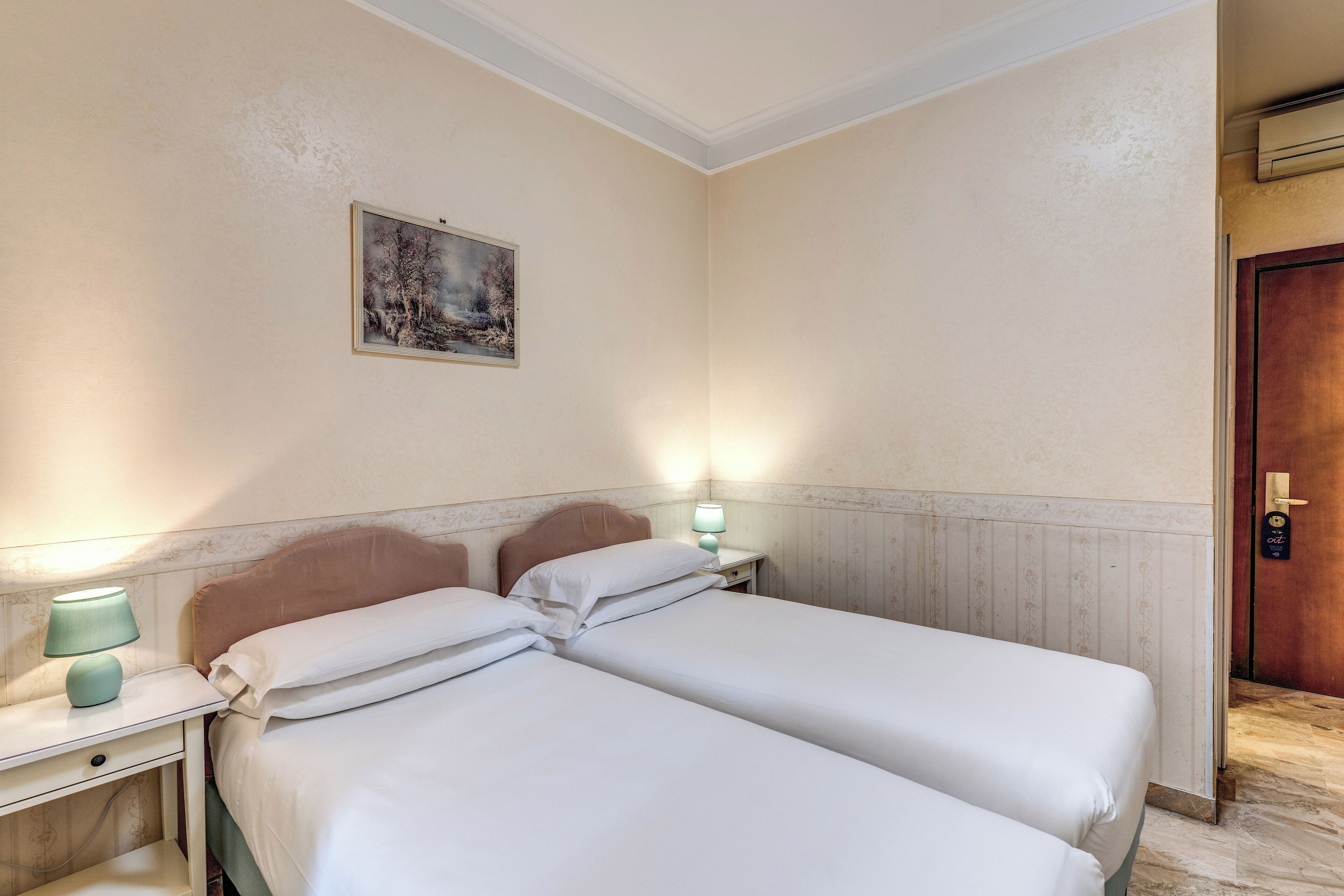 A hotel room with two beds, a nightstand with a lamp, a painting on the wall, and a wooden door.