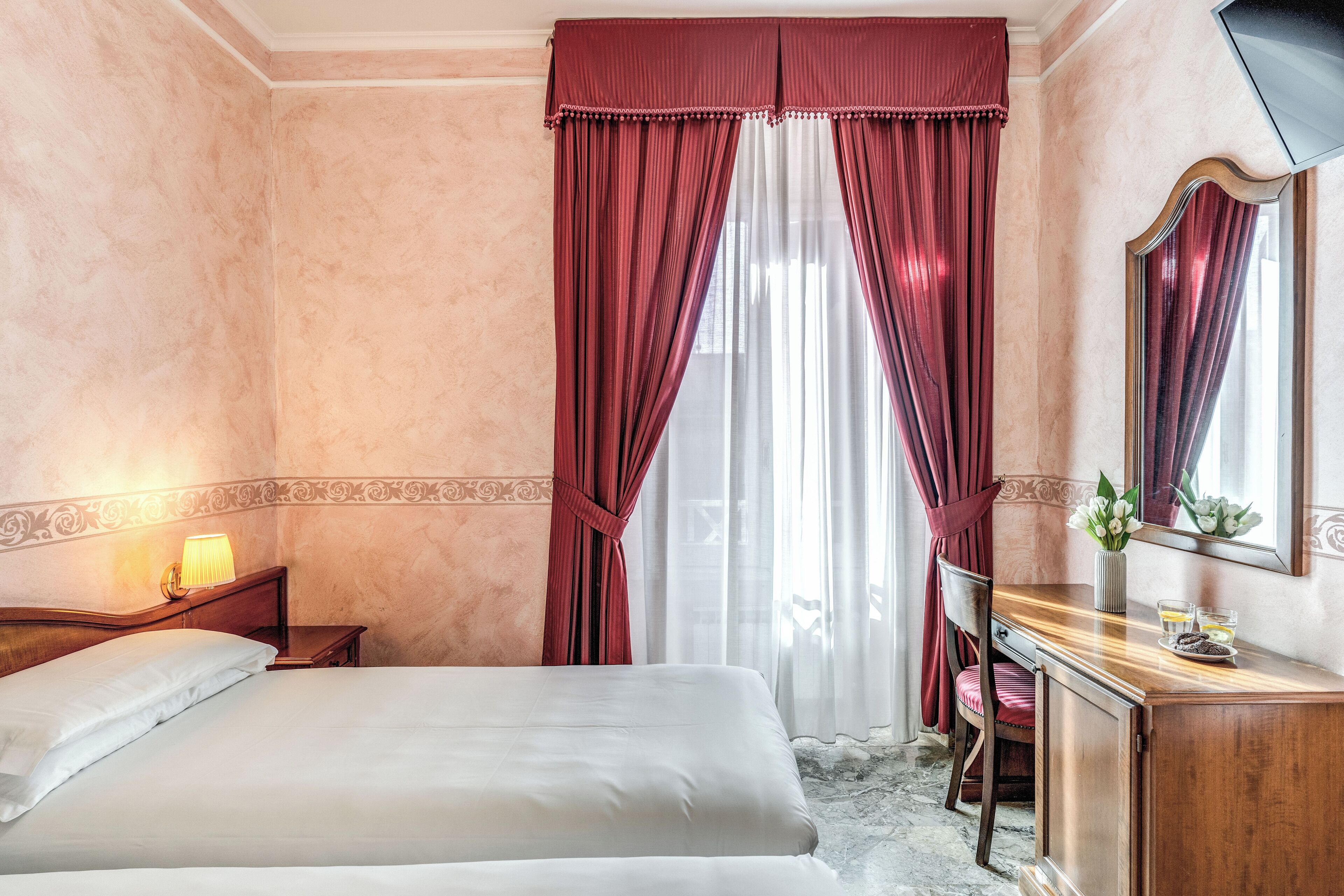 A hotel room with a bed, a desk with a mirror, a chair, a nightstand with a lamp, and a window with red curtains.