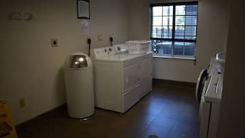 A laundry room with a front-loading washer and dryer, a trash can, and a window with blinds.