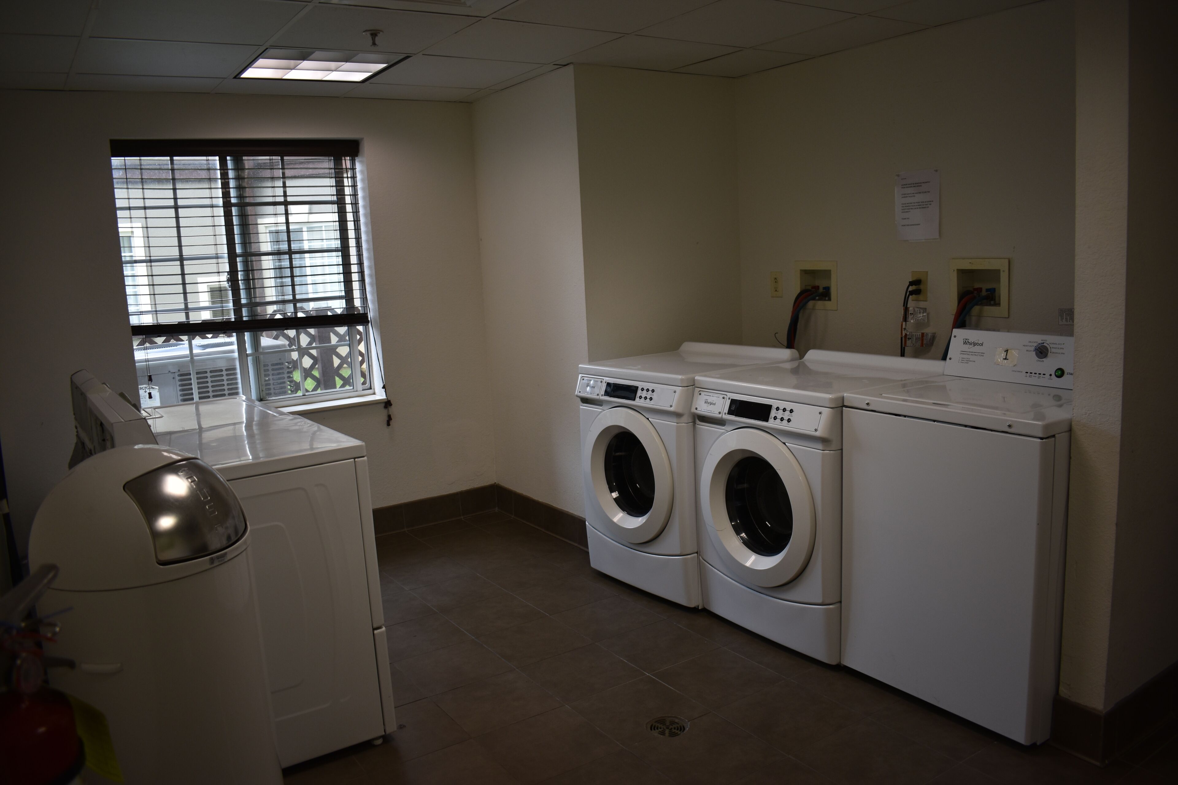A laundry room with two front-loading washers and dryers, a window with blinds, and a fire extinguisher on the left.