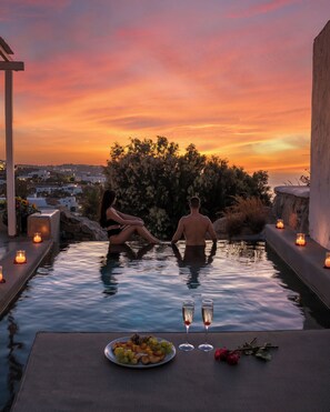 A couple relaxing in a private infinity pool with candles, surrounded by a scenic view of a sunset and a cityscape. - Ostraco Suites (Mykonos)