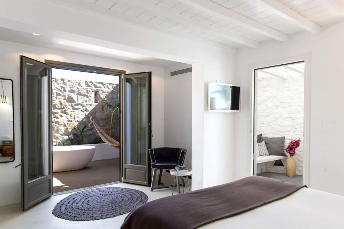 Un dormitorio moderno con una cama grande, una mesita redonda pequeña, una silla, un televisor y vistas a un muro de piedra exterior y una hamaca.