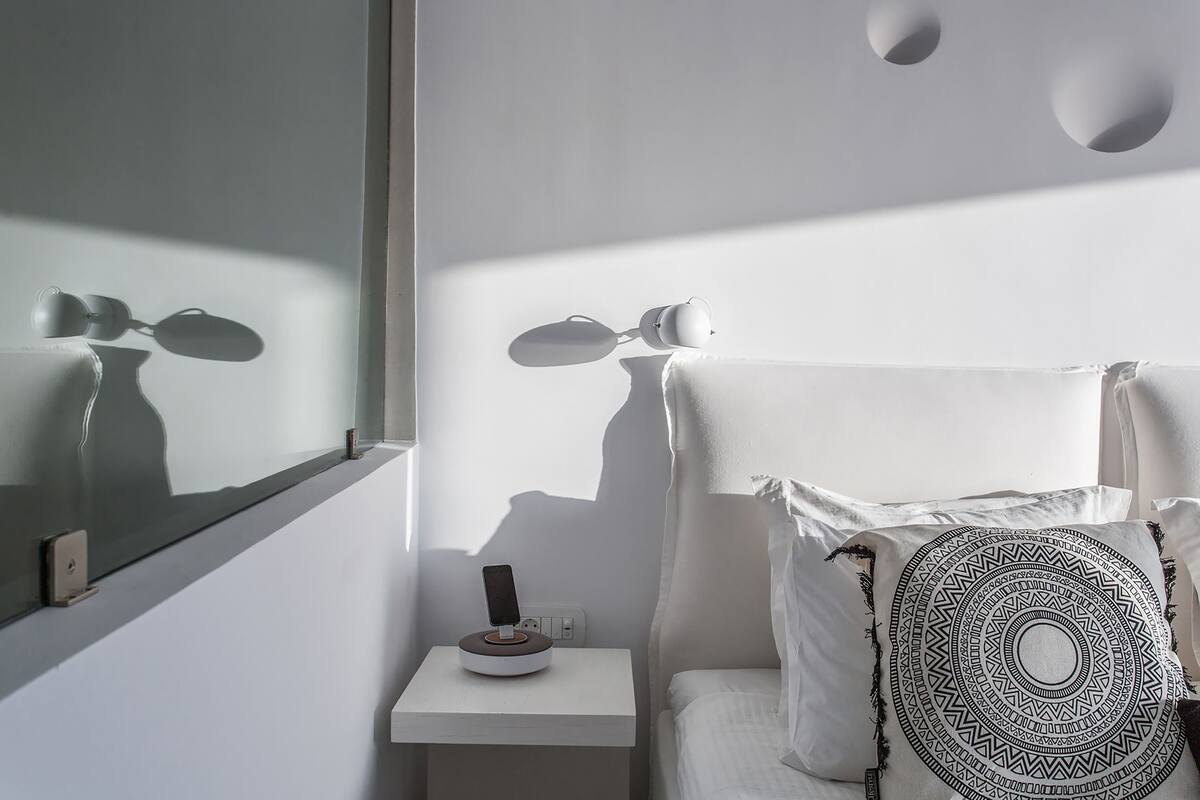 Una habitación minimalista con una mesita de noche, una estación de carga para teléfono inteligente, un cojín estampado y un espejo que refleja la luz del cuarto.