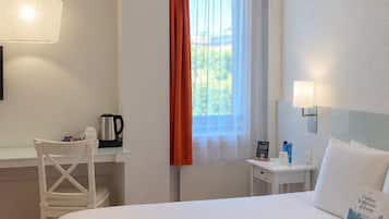Habitación de hotel con una cama grande, un escritorio con silla, una ventana con cortinas, una mesita de noche con lámpara y vistas al exterior con vegetación.