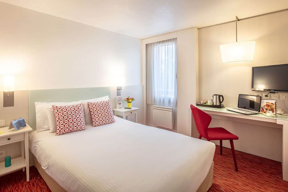 Habitación de hotel con una cama grande, un escritorio con computadora, una silla roja y un televisor.