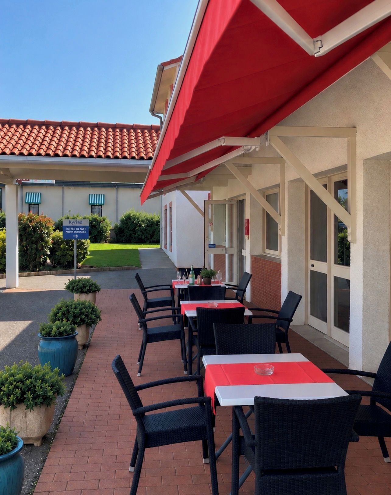 Un espace extérieur aménagé avec des tables et des chaises, un auvent rouge et une entrée de bâtiment avec une enseigne.