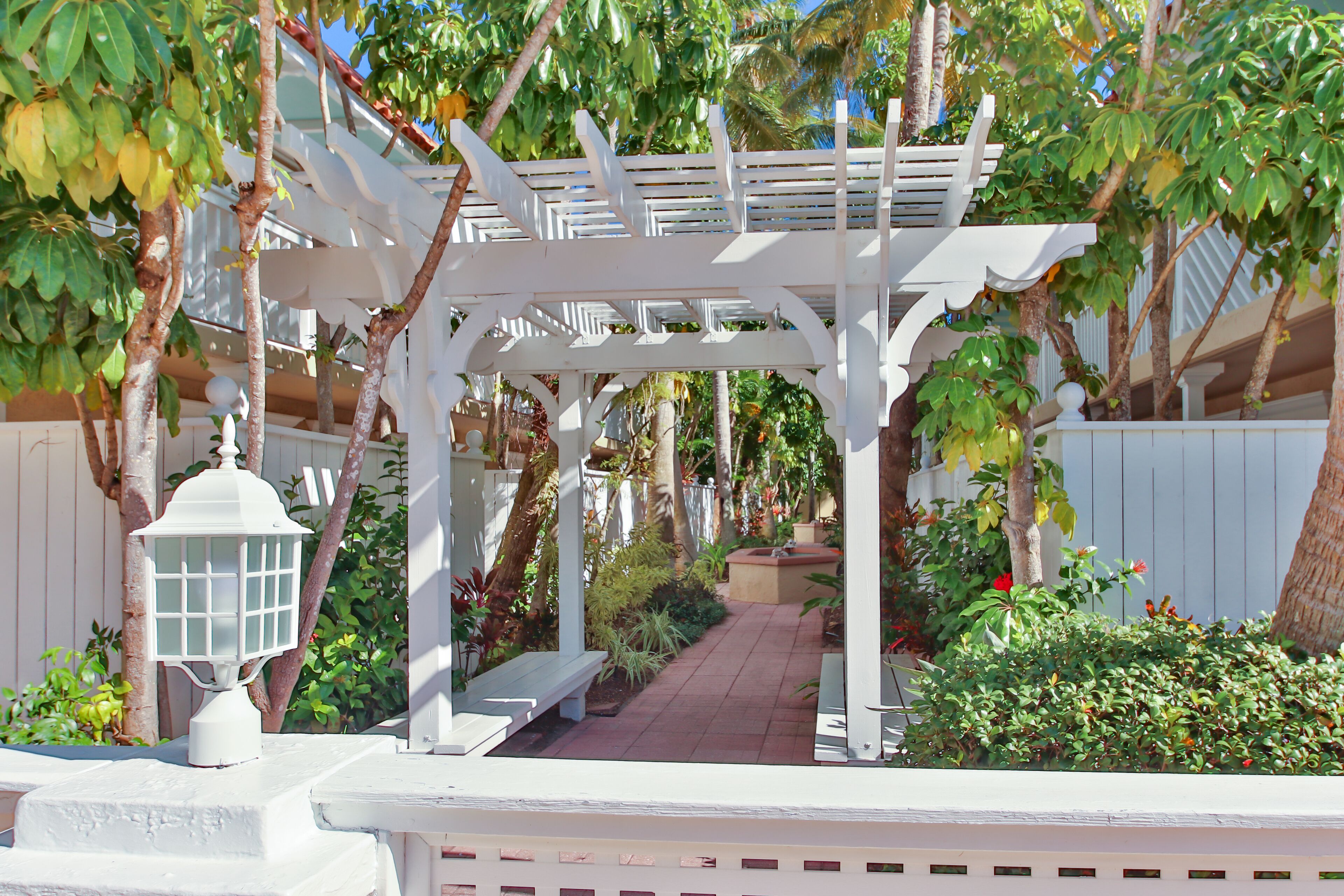A white pergola structure with a bench under it, surrounded by lush greenery and a pathway.
