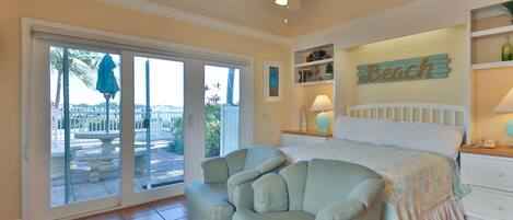 A bedroom with a bed, a ceiling fan, a sliding glass door to a patio, and a 'Beach' sign on the wall.