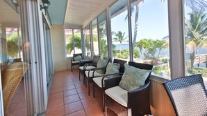A covered outdoor seating area with wicker chairs and cushions, large windows offering a view of palm trees and a dock.