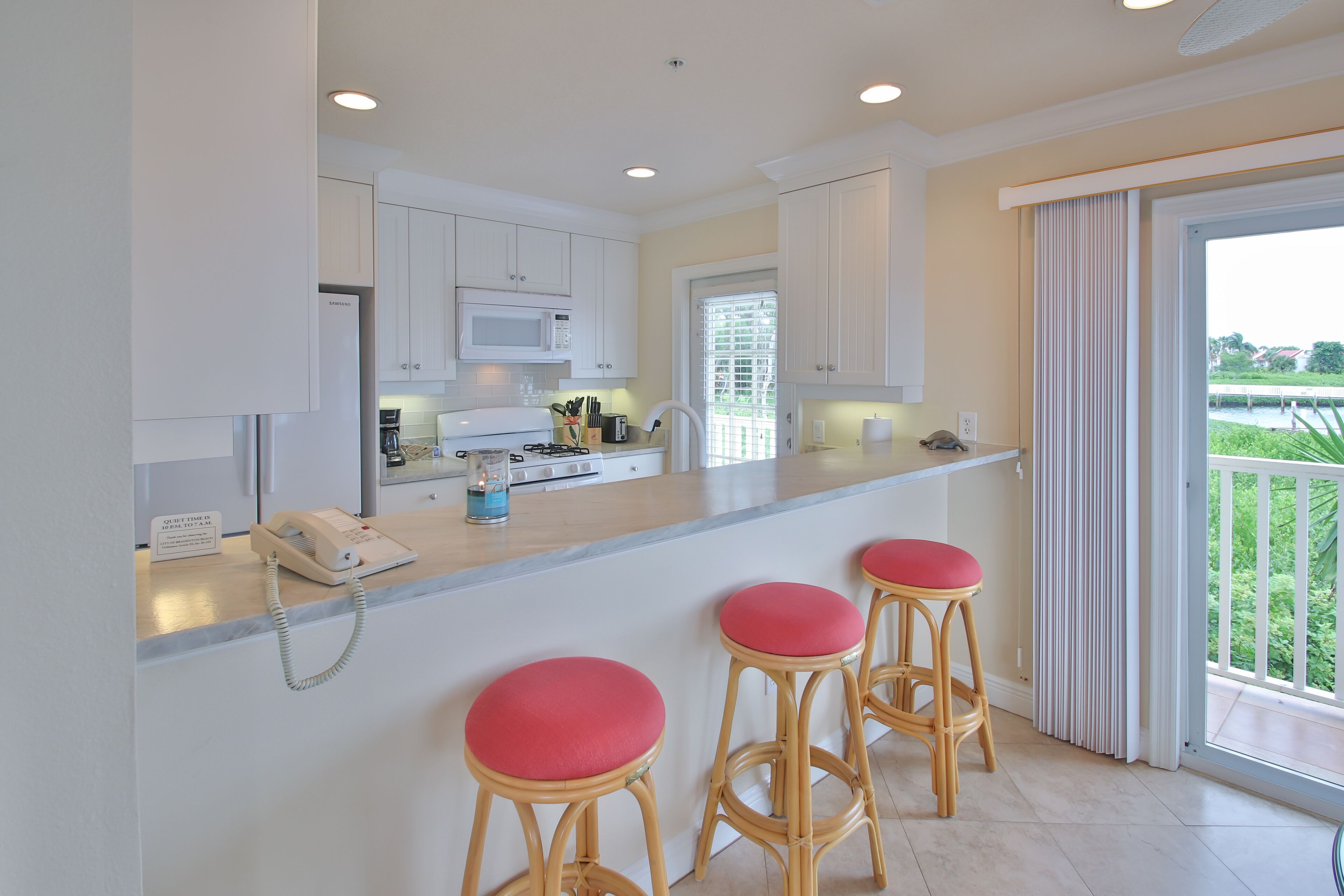 A modern kitchen with white cabinets, a microwave, and a gas stove. There are three wooden stools with red cushions. A phone is on the counter, and a window with blinds is visible.
