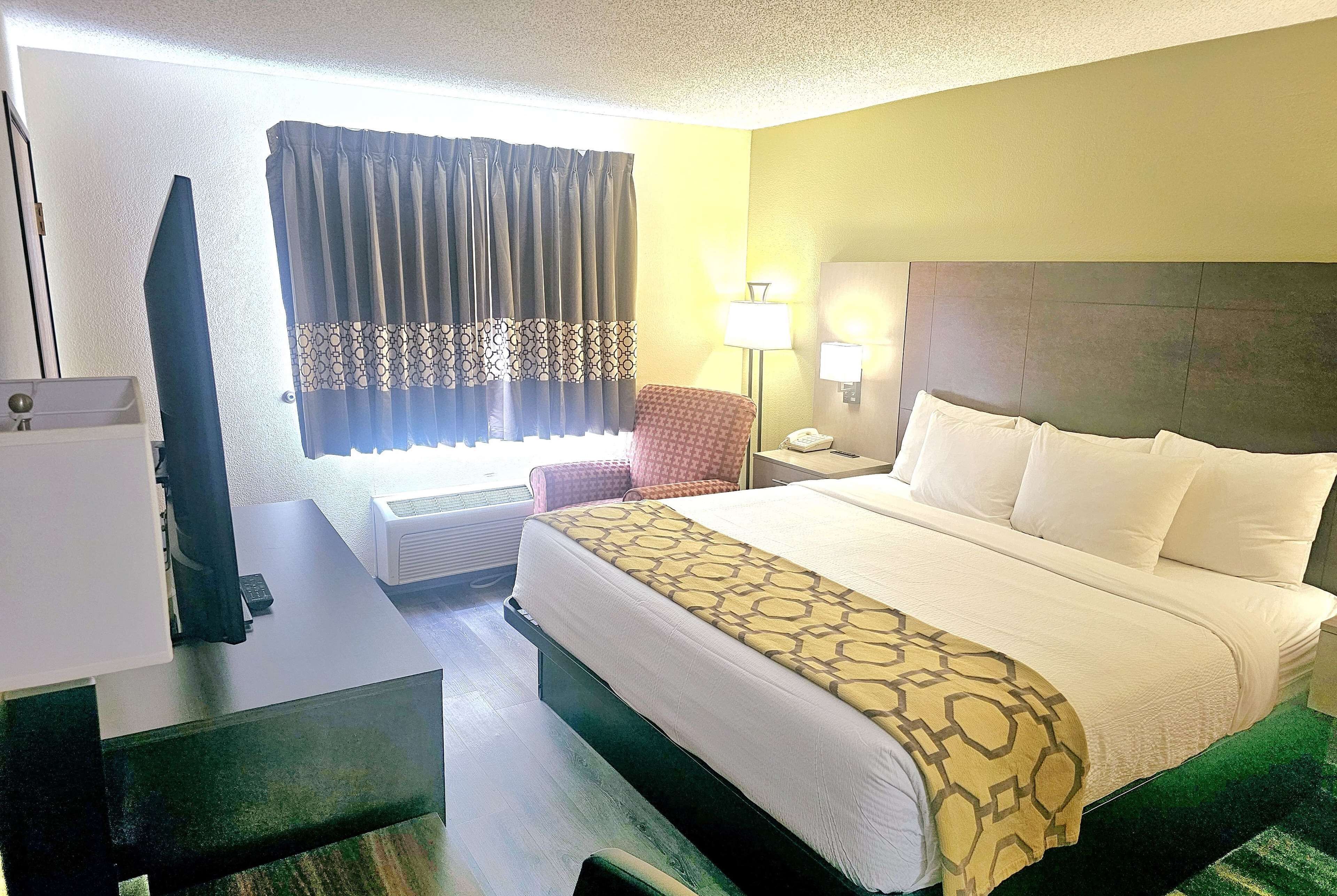 Une chambre d’hôtel avec un lit, un bureau, une chaise, une lampe et une fenêtre avec des rideaux.