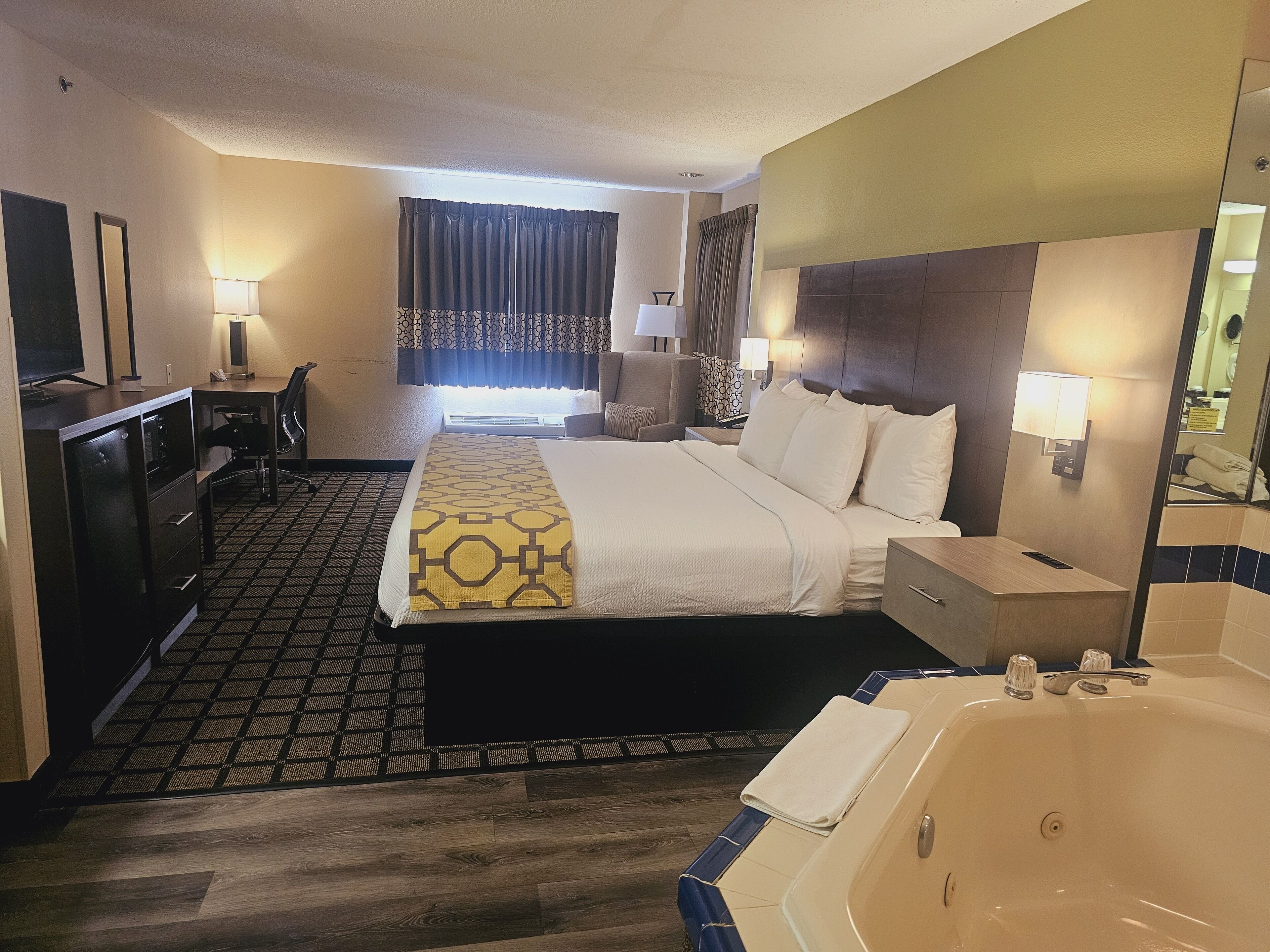 Habitación de hotel con cama, escritorio, silla, lámpara de escritorio, mesita de noche, lámpara, baño con bañera y espejo.