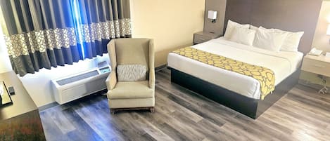 A hotel room with a bed, a chair, a bedside table, a nightstand, a wall-mounted lamp, and a window with curtains.