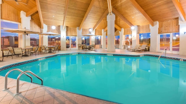 An indoor swimming pool with clear blue water, surrounded by wooden beams and columns, with seating areas and large windows. - Baymont by Wyndham Rolla (Rolla)