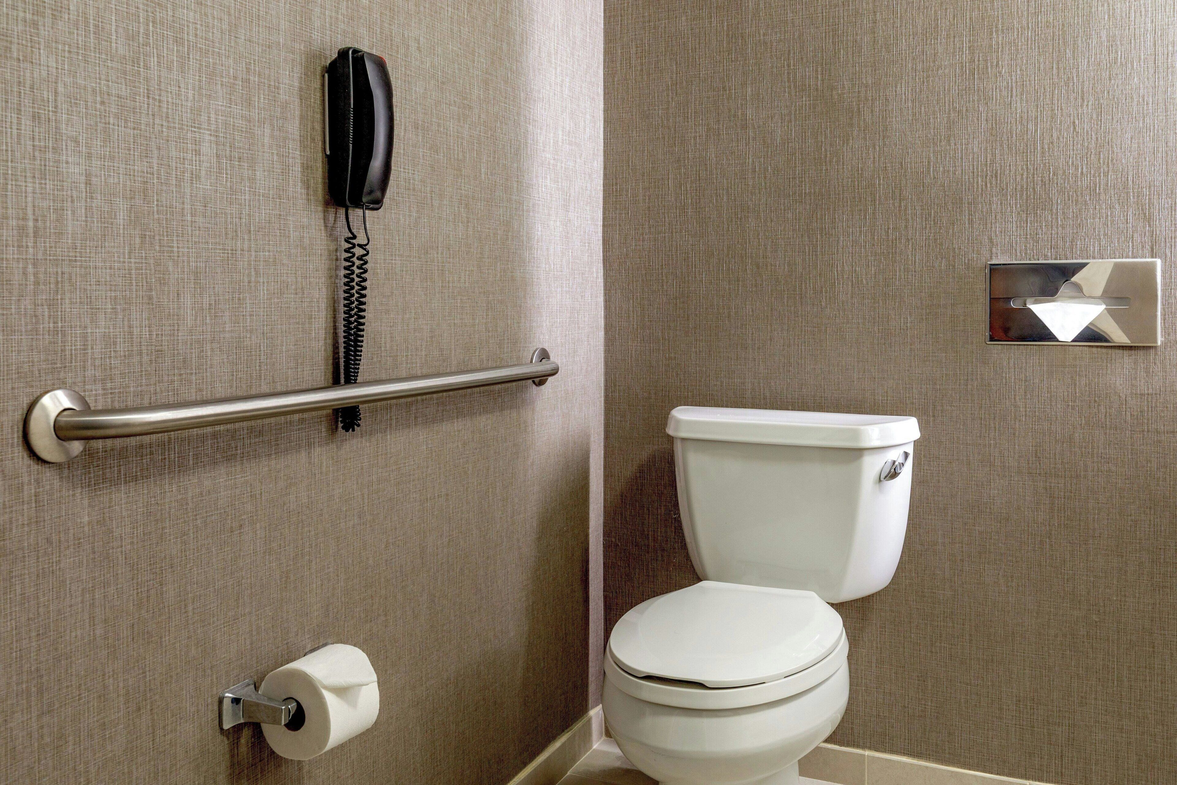 A bathroom with a toilet, a handrail, a toilet paper holder, and a telephone mounted on the wall.