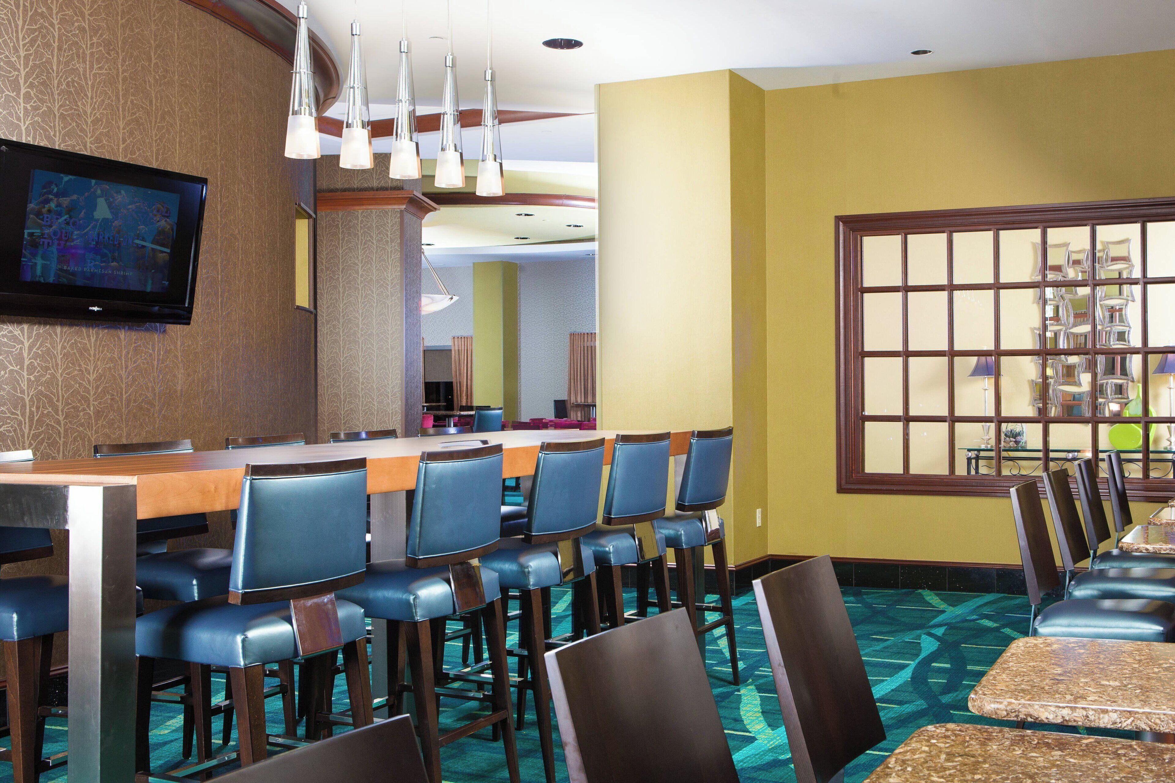 A hotel bar with multiple seating options, including bar stools and tables, a television mounted on the wall, and a large window with a view of the interior.
