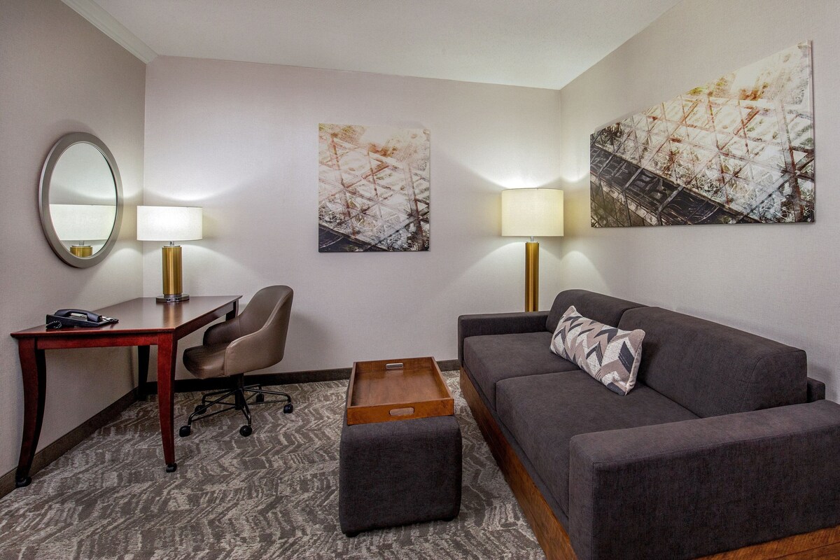 A hotel room with a gray sofa, a wooden side table, a desk with a lamp, a chair, and two wall-mounted lamps.