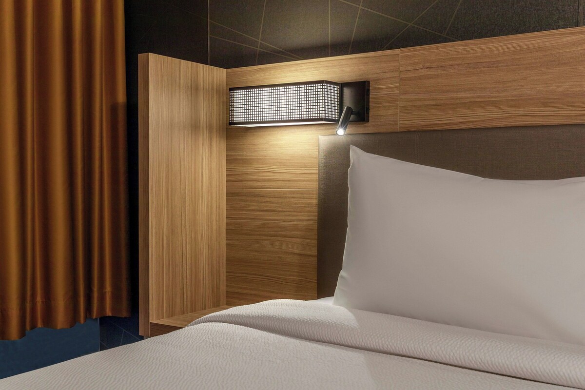 A hotel room with a bed, a headboard, a wall-mounted light fixture, and a curtain.