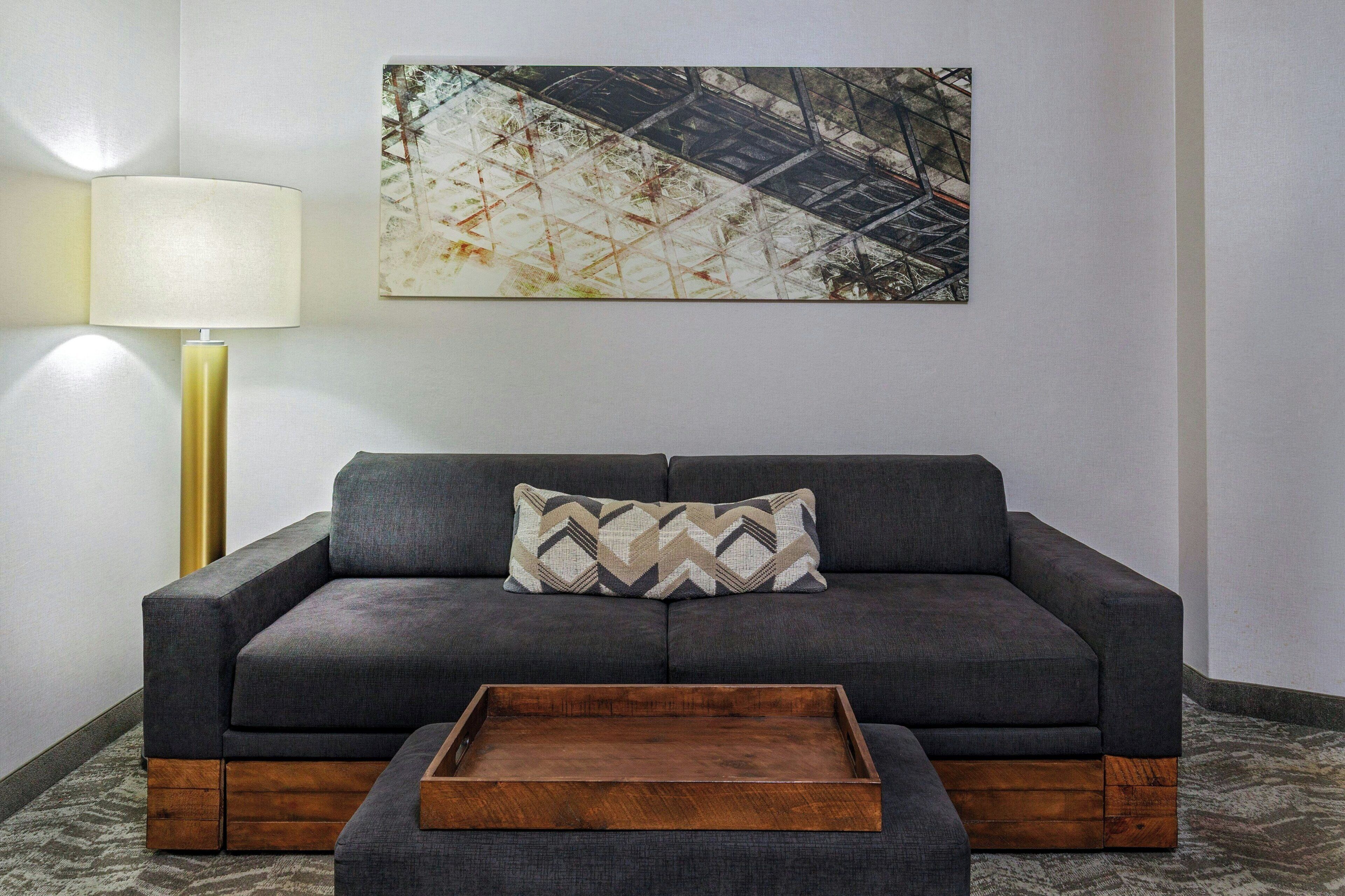 A grey sofa with a patterned pillow, a wooden tray, and a lamp on a patterned rug.
