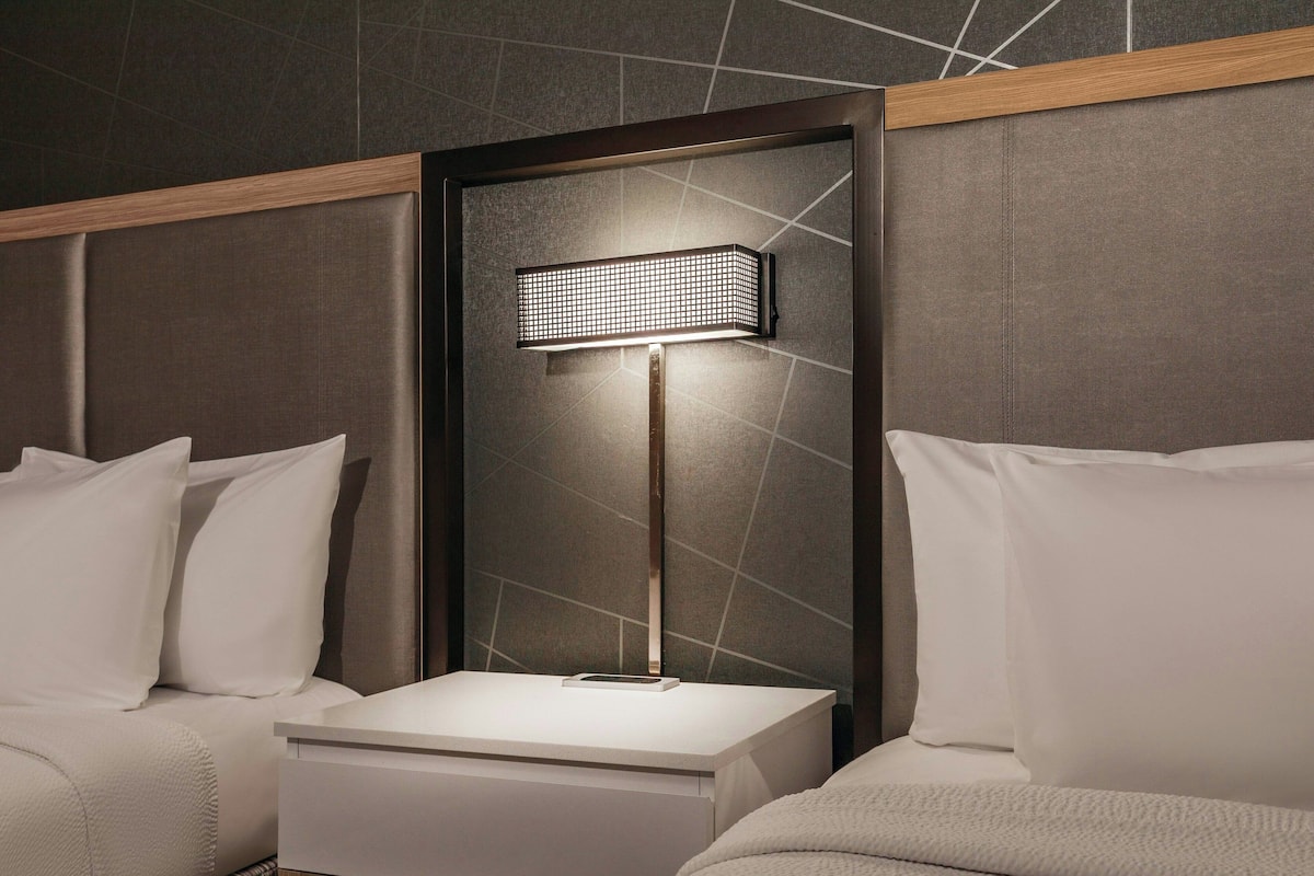 A modern hotel room with a bed, a bedside table, and a wall-mounted lamp.
