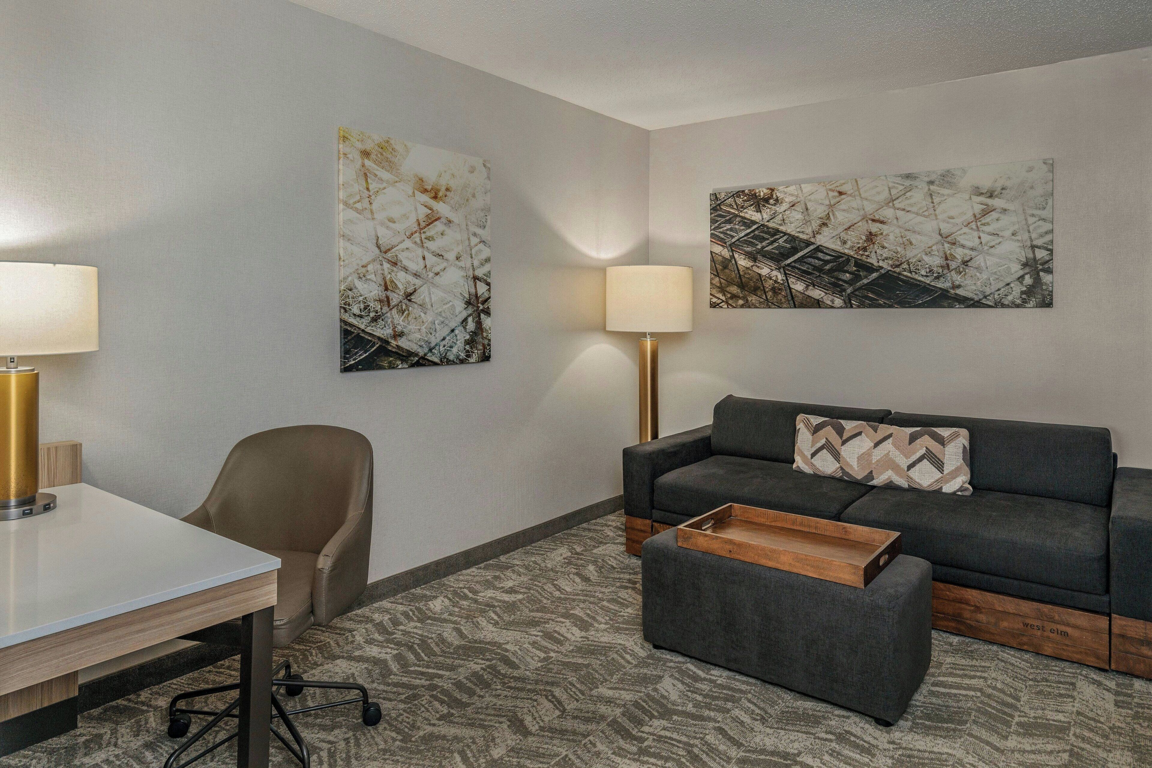 A modern hotel room with a desk, office chair, sofa, ottoman, two wall-mounted lamps, and abstract art on the walls.