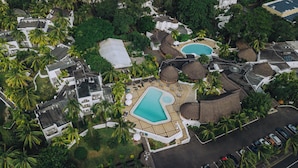 Vista aérea de un resort con dos piscinas, palmeras y una estructura de techo de paja.
