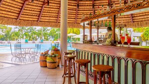 Una zona de bar con techo de paja, taburetes de madera y un área de estar junto a la piscina con sombrillas.