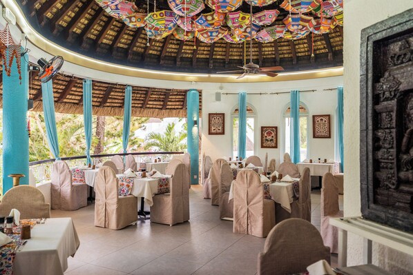 Un restaurante con mesas y sillas redondas, techo de paja y elementos decorativos colgantes.