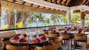 Un restaurante con mesas dispuestas para comer, con vistas a una playa de palmeras y el océano.