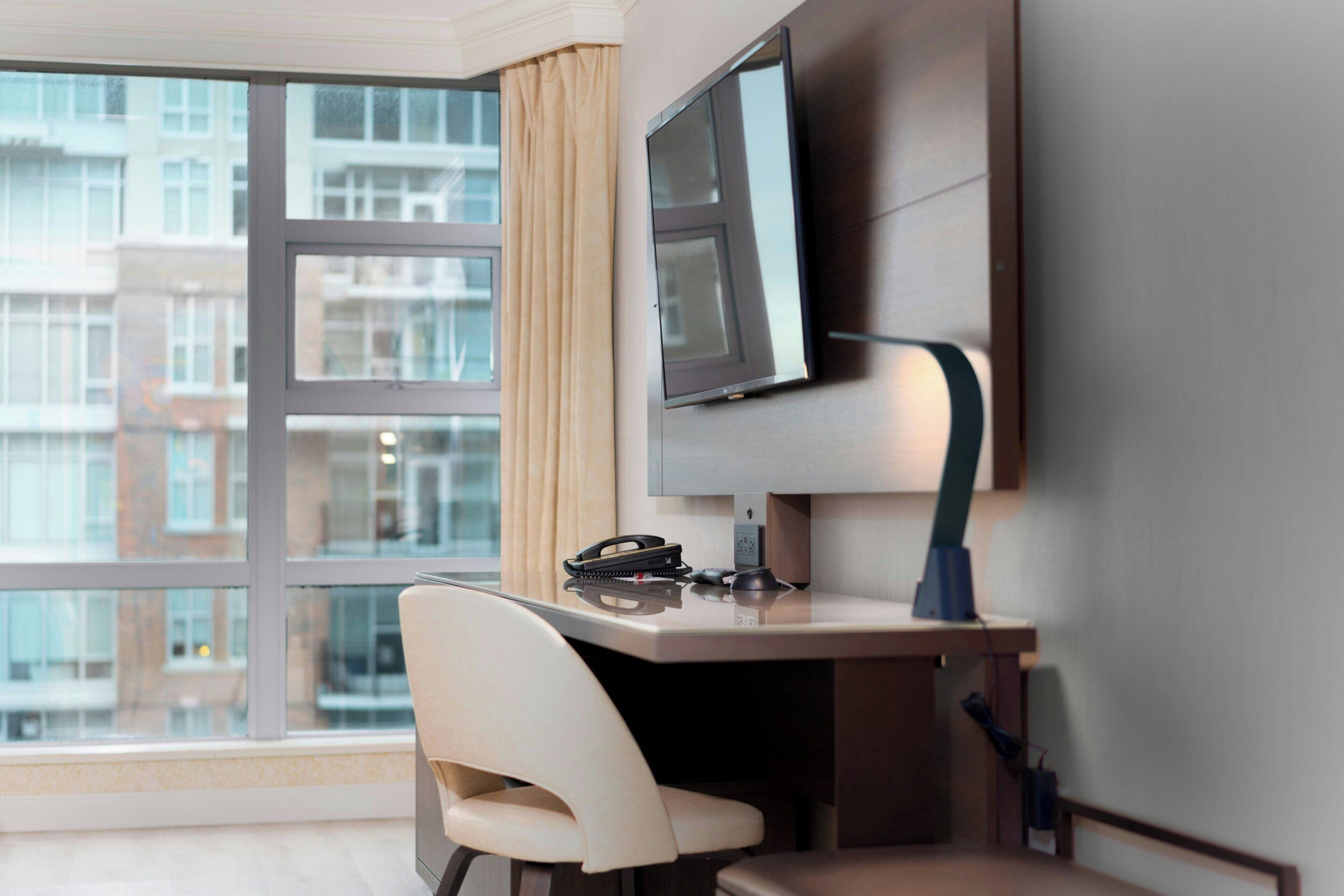 A modern hotel room with a large window, a desk with a flat-screen TV, a chair, and a telephone.