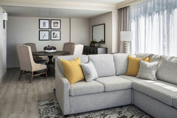 Una sala de estar moderna con un sofá seccional gris, dos sillones, una mesa de comedor redonda y una mesa auxiliar. Hay cuadros enmarcados en la pared, un espejo sobre el sofá y una lámpara junto a la ventana.