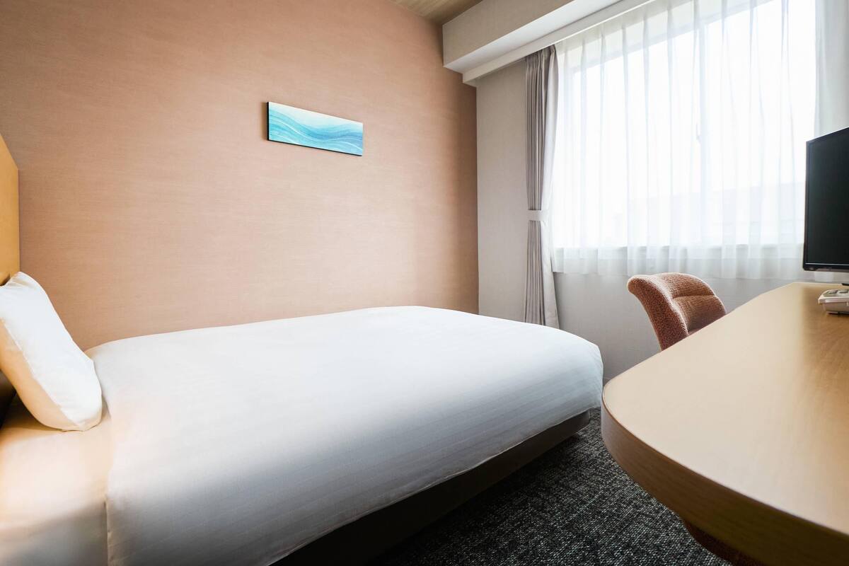 A hotel room with a bed, a desk, a chair, a television, a painting on the wall, and a window with curtains.