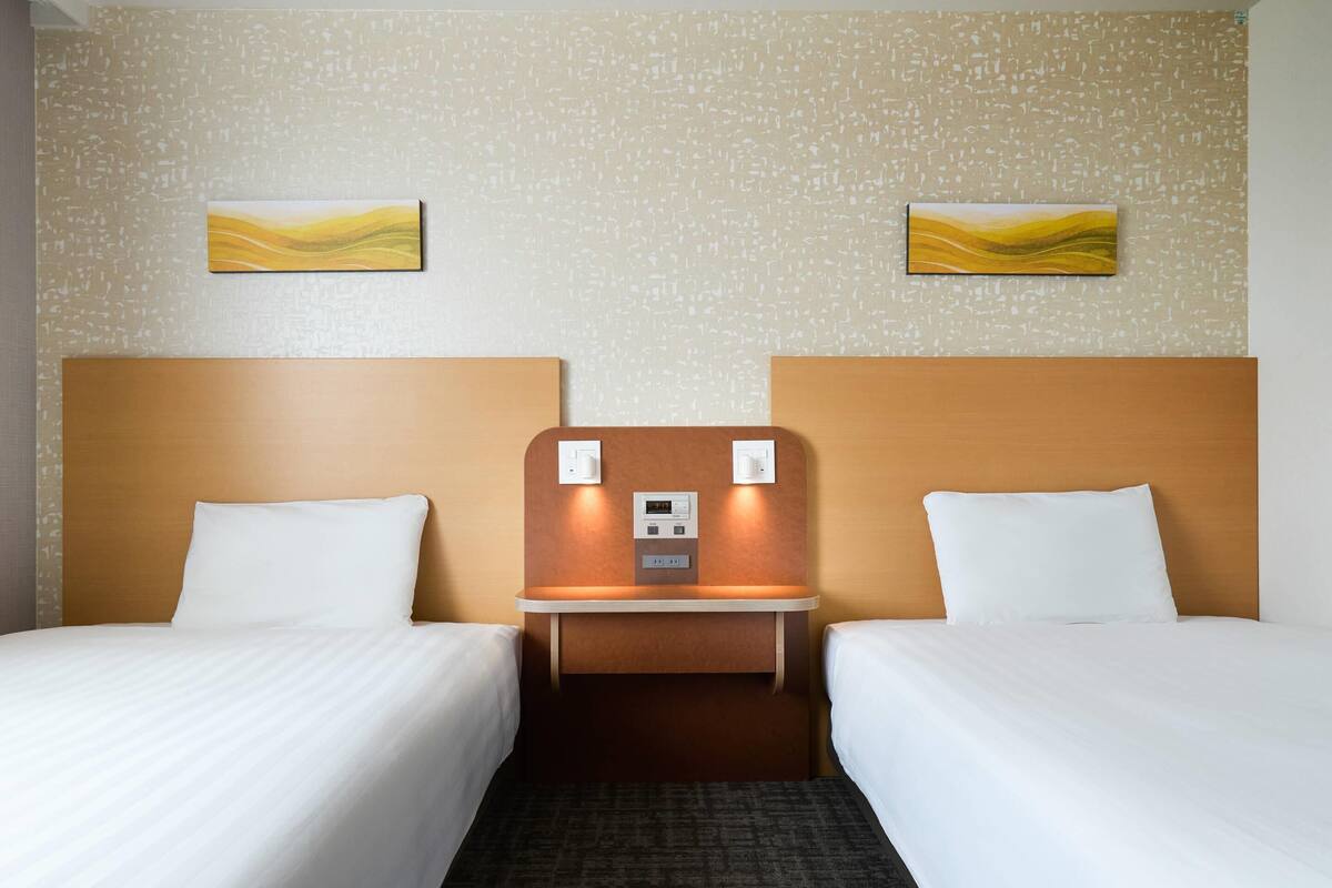 A hotel room with two single beds, a small bedside table with a control panel, and a textured wall with abstract artwork.