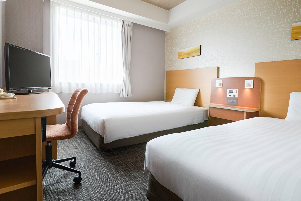 A hotel room with two beds, a desk with a TV, a chair, and a bedside table with a lamp.