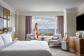 A hotel room with a large bed, two armchairs, a small table, a TV, and a view of mountains.