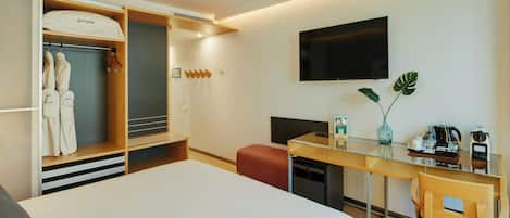 Habitación de hotel con cama, escritorio y silla, televisión, minibar, planta y armario con batas.