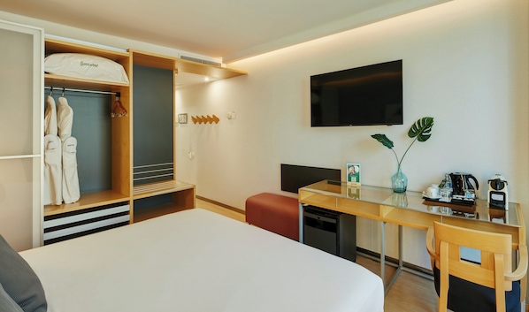 Habitación de hotel con cama, escritorio y silla, televisión, minibar, planta y armario con batas.