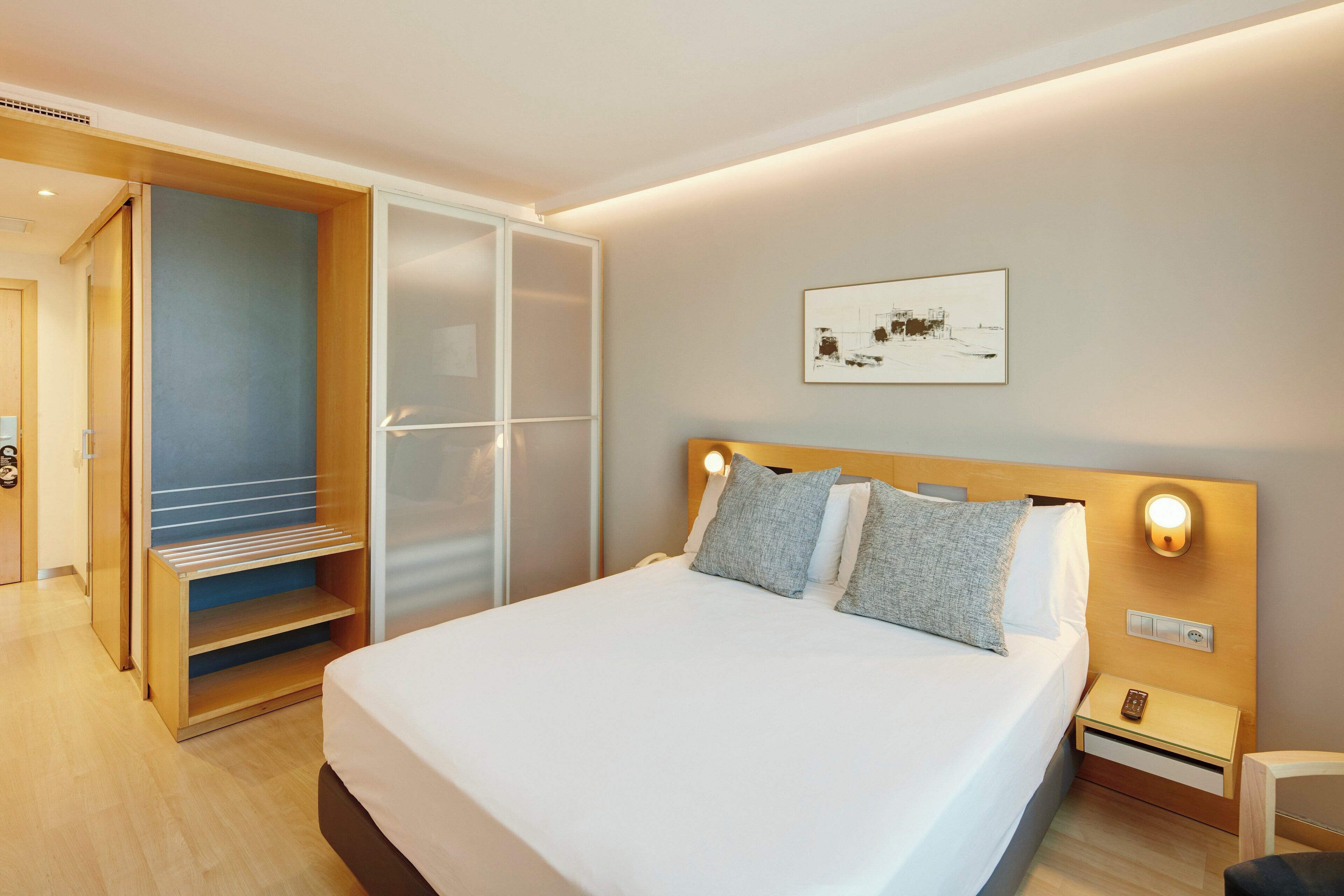A hotel room with a bed, a nightstand, a wall-mounted light, a door, and a framed picture on the wall.