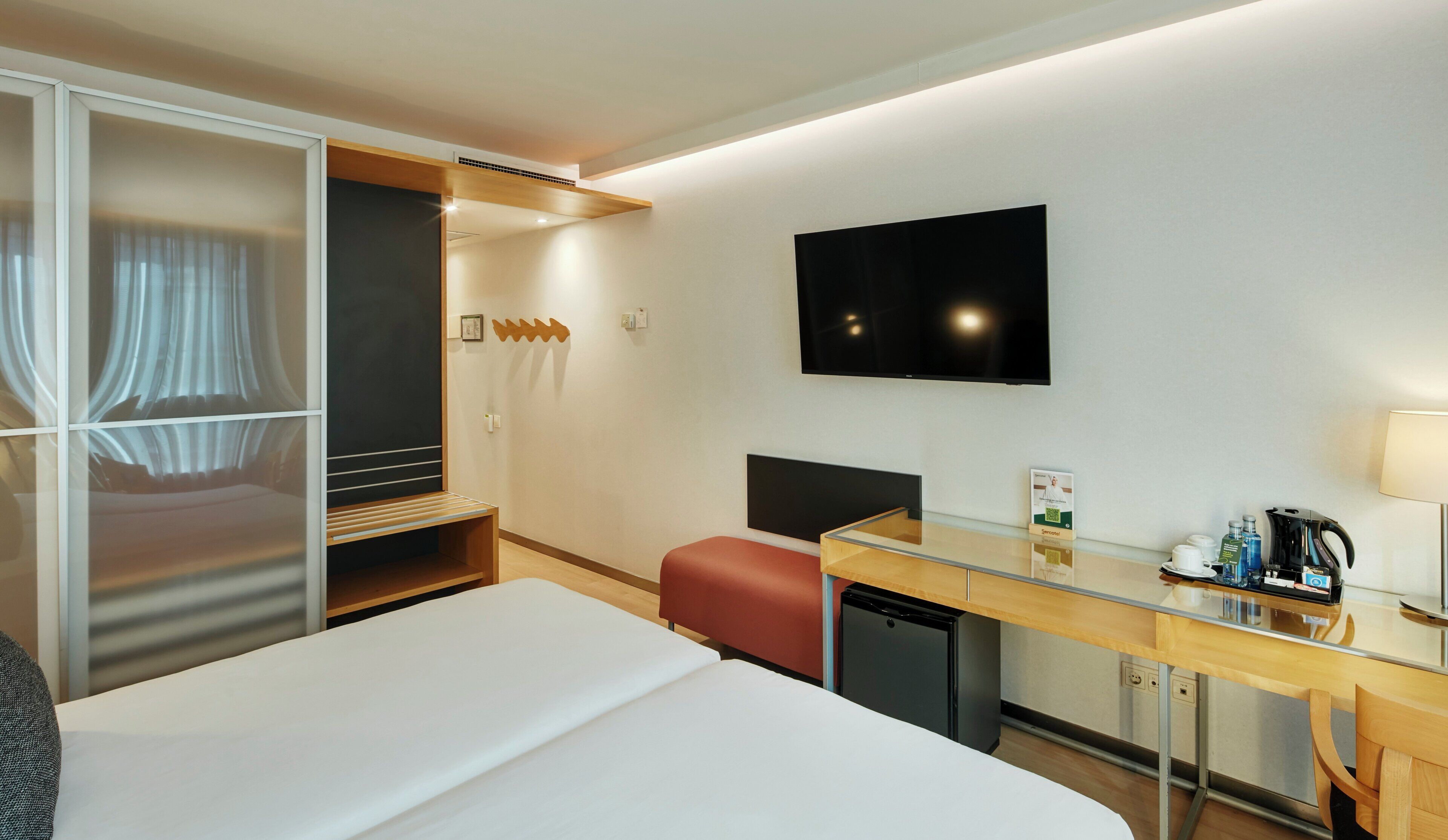 A hotel room with a bed, a desk with a computer, a TV, a mini-fridge, and a wardrobe.