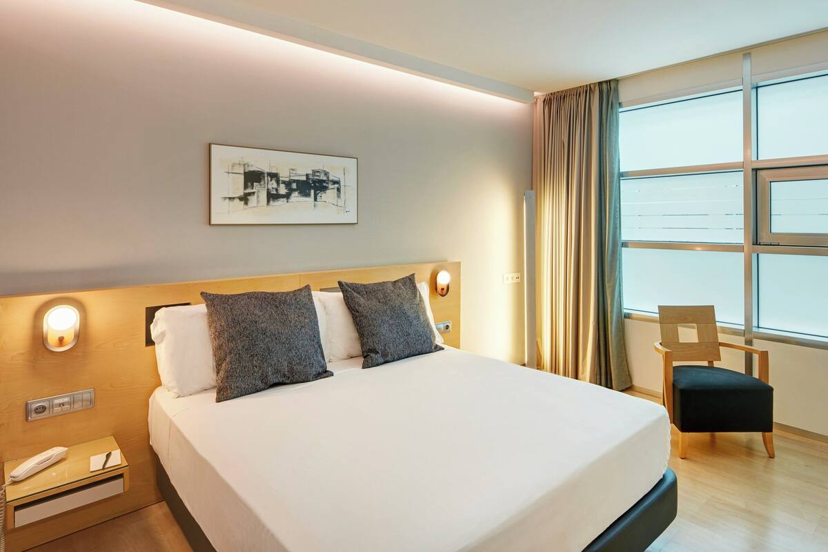 Una habitación de hotel con una cama, una silla, una mesita de noche con teléfono, una lámpara de pared y un ventanal con cortinas.