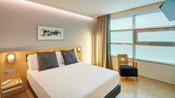 A hotel room with a bed, a chair, a bedside table with a phone, a wall-mounted light, and a window with curtains.