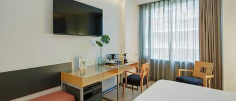 Quarto de hotel com cama, escrivaninha, cadeira, televisão e janela com cortinas.