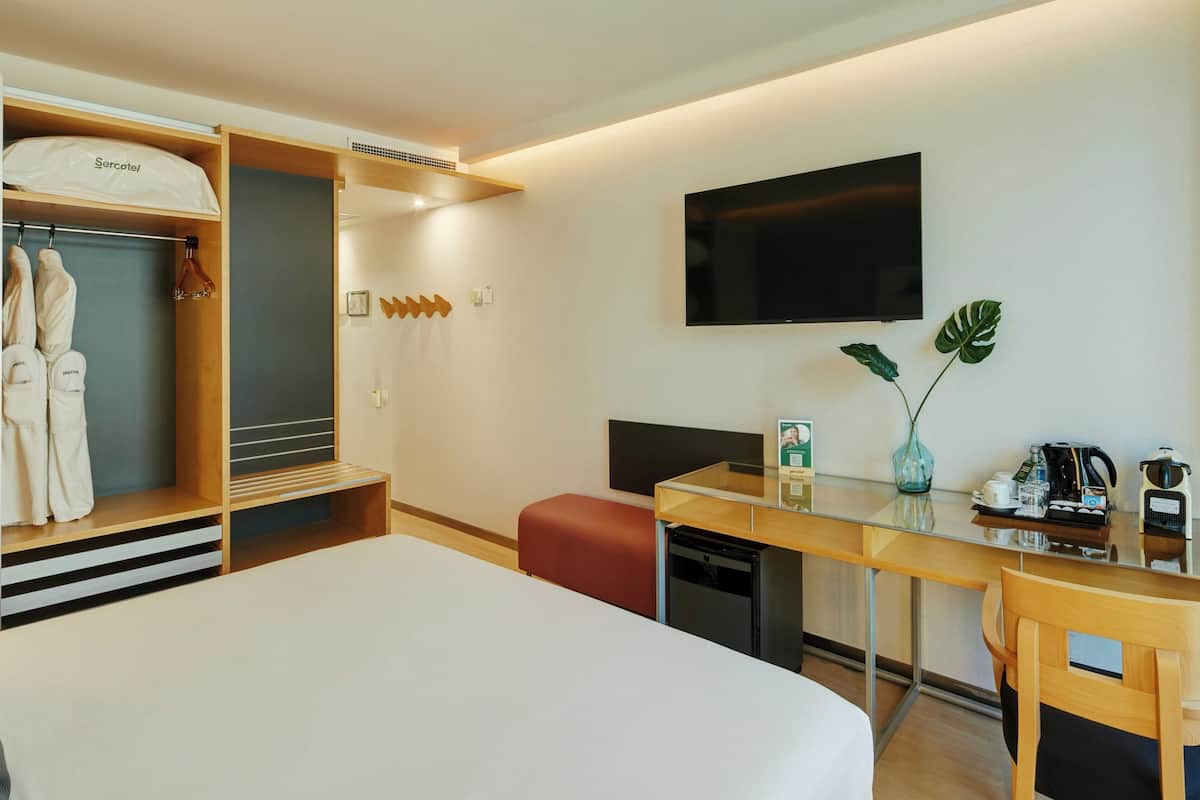 Une chambre d’hôtel comprenant un lit, un bureau avec une chaise, une télévision, un minibar, une plante et une armoire avec des peignoirs.