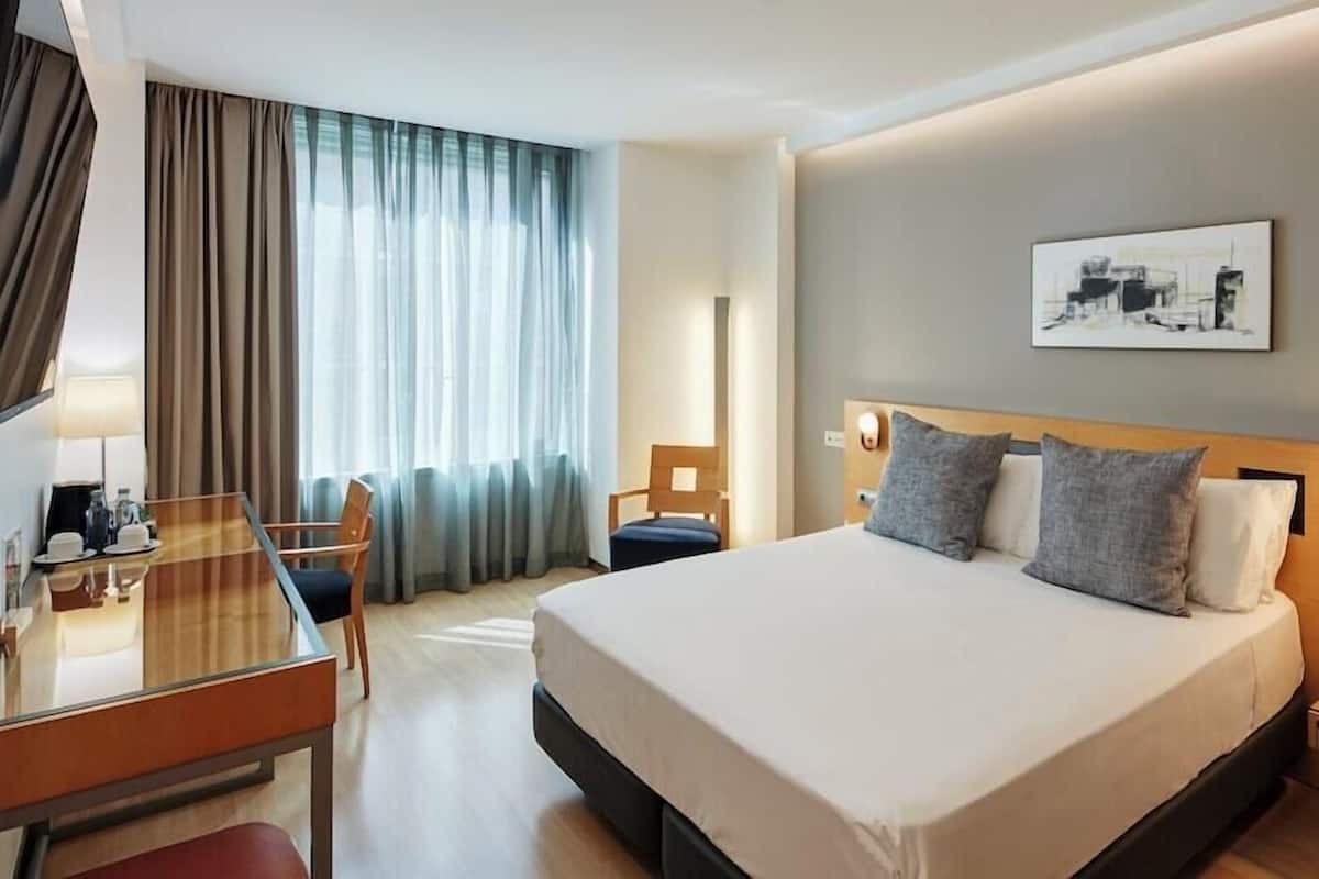 Une chambre d’hôtel comprenant un lit, des tables de chevet, un bureau, une chaise et une fenêtre avec des rideaux.