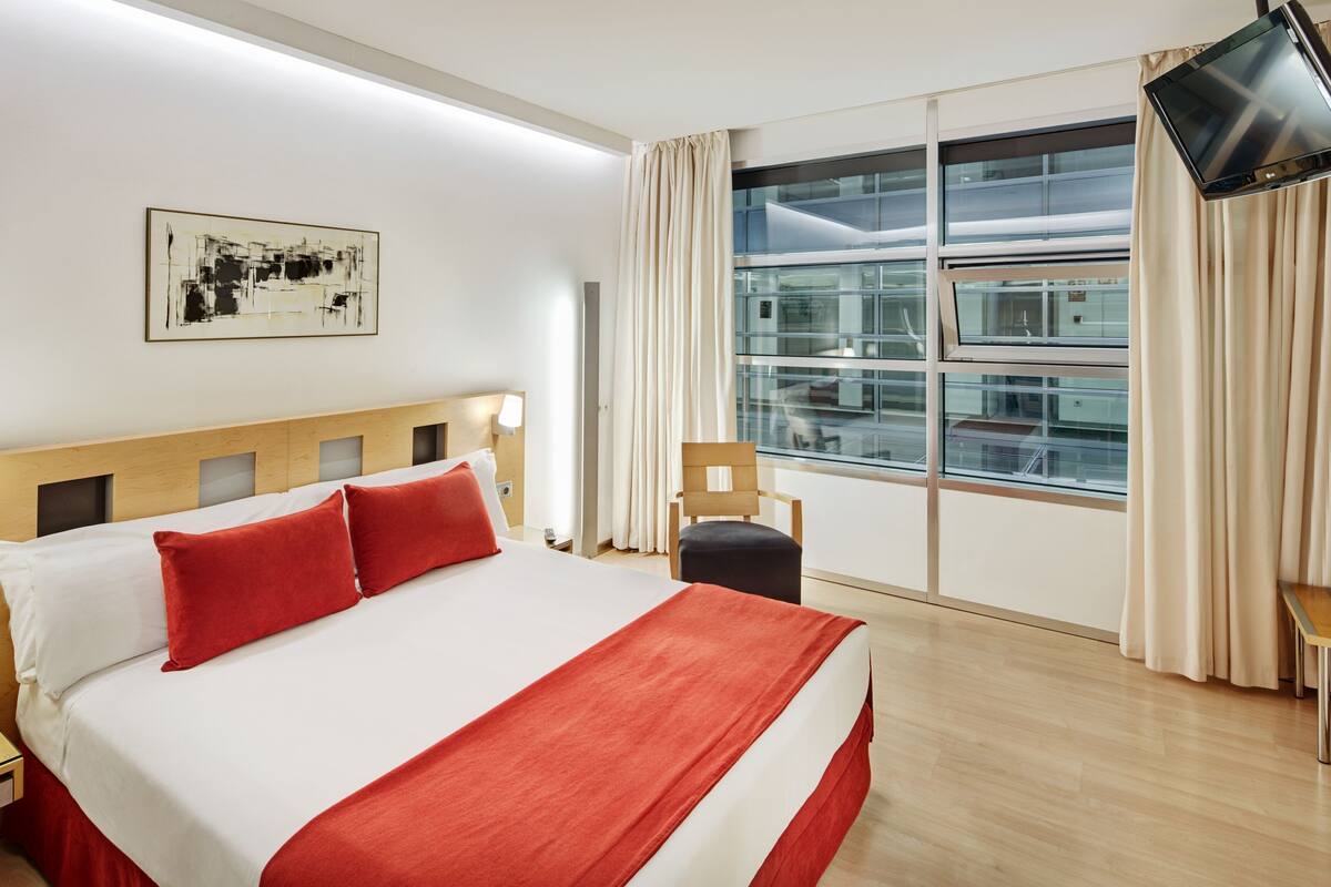 Une chambre d’hôtel avec un grand lit, une télévision, une fenêtre avec des rideaux et un tableau au mur.