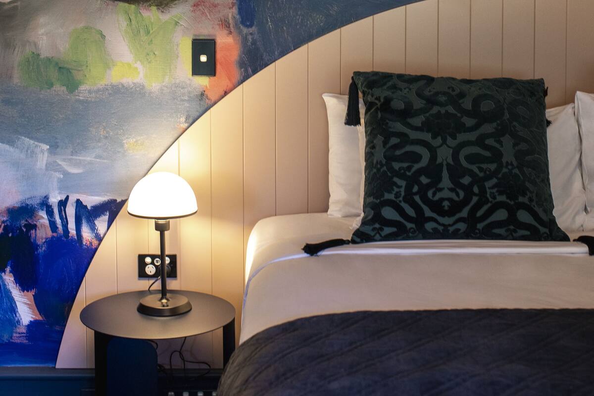 A hotel room with a large abstract painting on the wall, a round bedside table with a lamp, and a bed with patterned pillows.