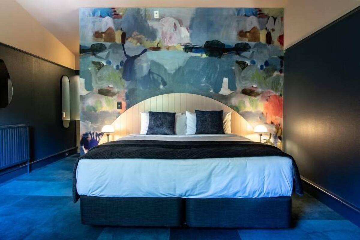 A modern hotel room with a large bed, a unique abstract mural on the wall, and two bedside lamps.