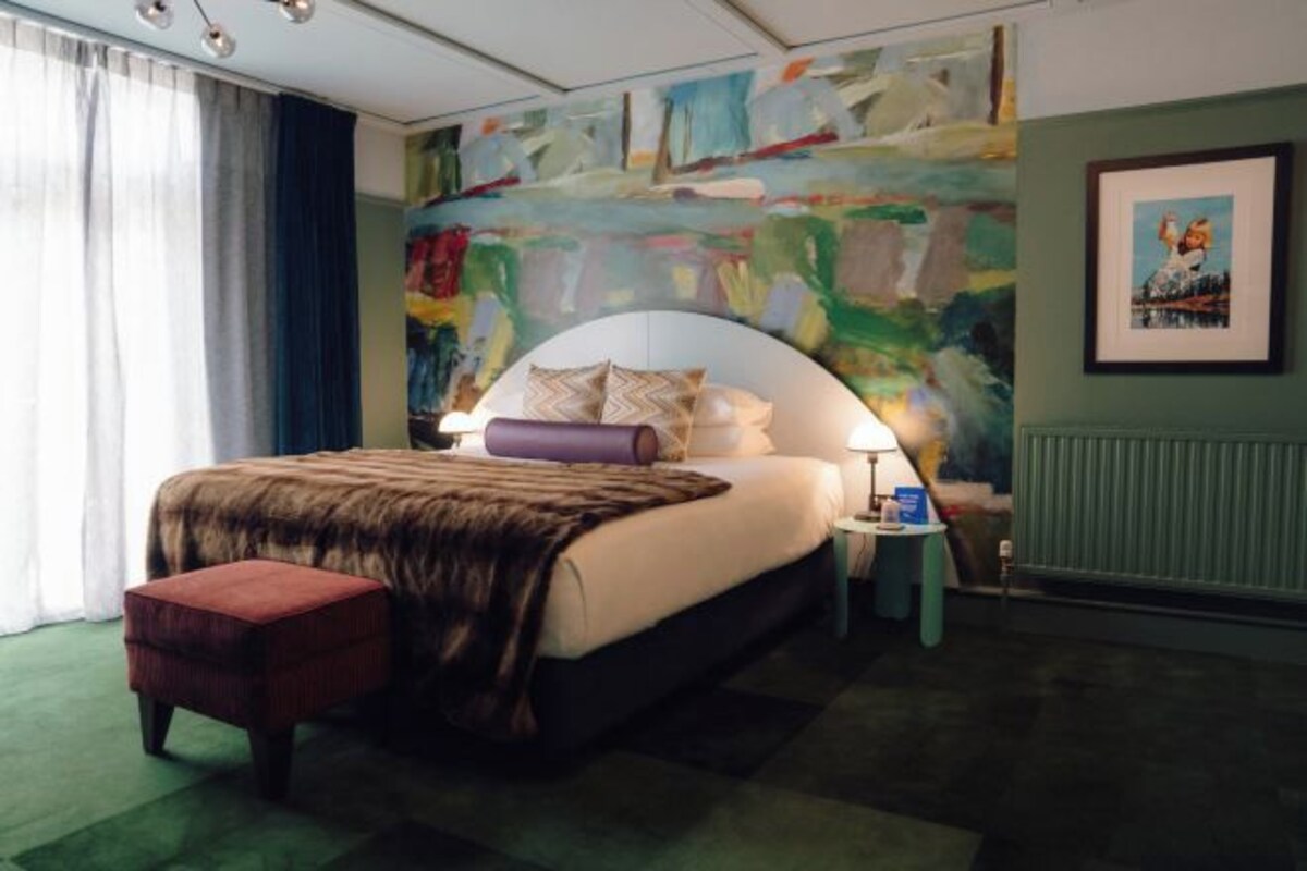 A bedroom with a curved headboard, a bedside table with a lamp, a red ottoman, and a large abstract painting on the wall.