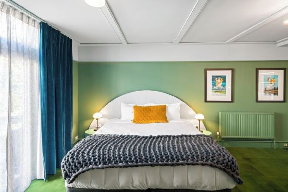 A neatly arranged bedroom with a bed, two bedside lamps, a radiator, and framed pictures on the wall.