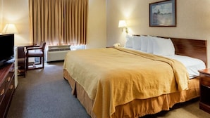 A hotel room with a bed, a desk with a TV, a lamp, and an air conditioning unit.